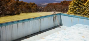 An empty and weathered pool with peeling surfaces, overlooking scenic mountains and trees, awaiting maintenance like fiberglass pool resurfacing.