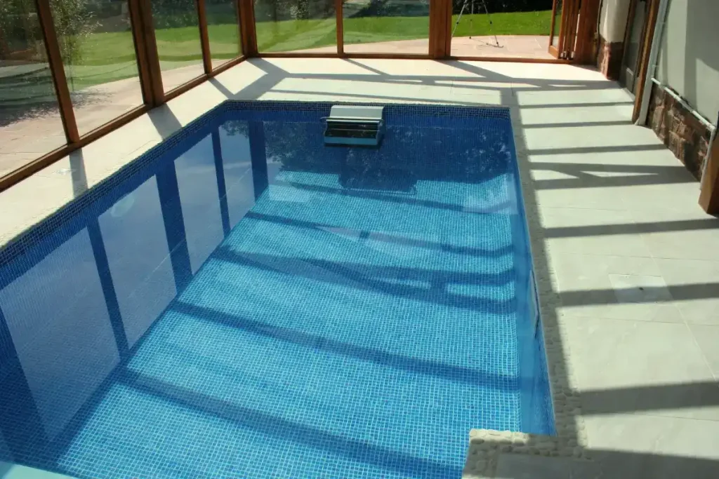 A compact fiberglass pool sizes with blue mosaic tiles, enclosed by glass walls with views of a lush garden.