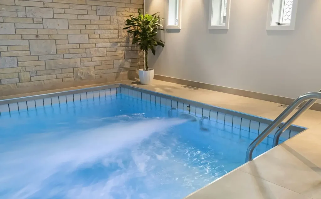 An indoor fiberglass pool above ground featuring hydrotherapy jets, surrounded by a sleek tile finish and decorative stone wall.