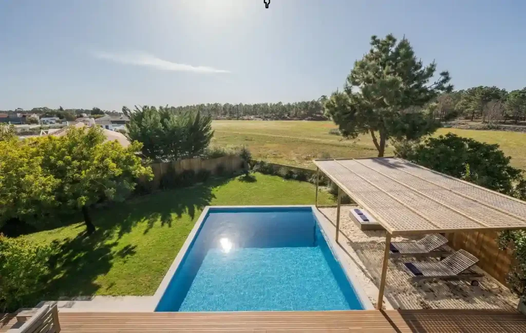 A modern backyard featuring an above-ground fiberglass pool surrounded by greenery and a wooden deck with a shaded sitting area.