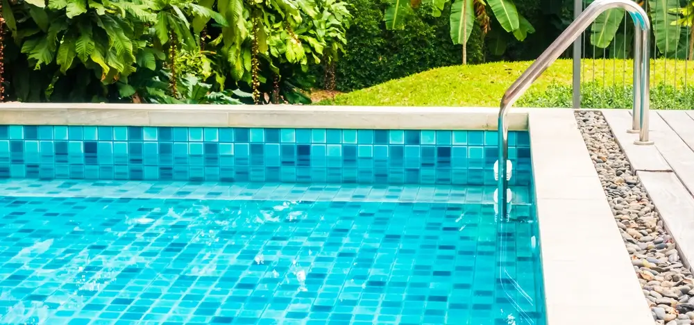 A fiberglass pool with vibrant blue tiles, surrounded by lush tropical greenery and a clean stone deck.