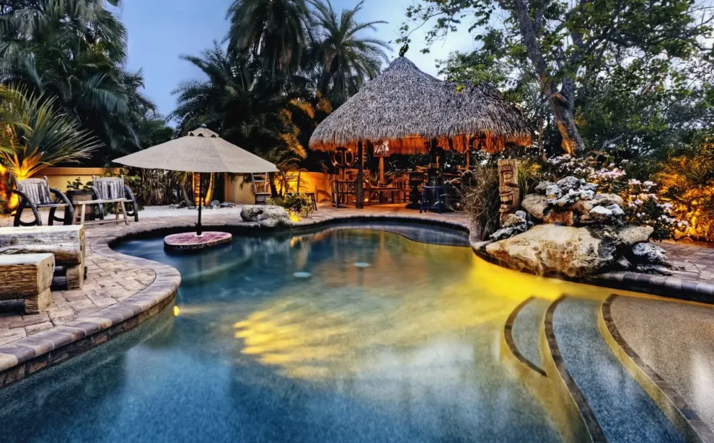 A uniquely shaped fiberglass pool for sale, illuminated at night, surrounded by tropical trees and a thatched-roof tiki bar, creating a resort-like ambiance.