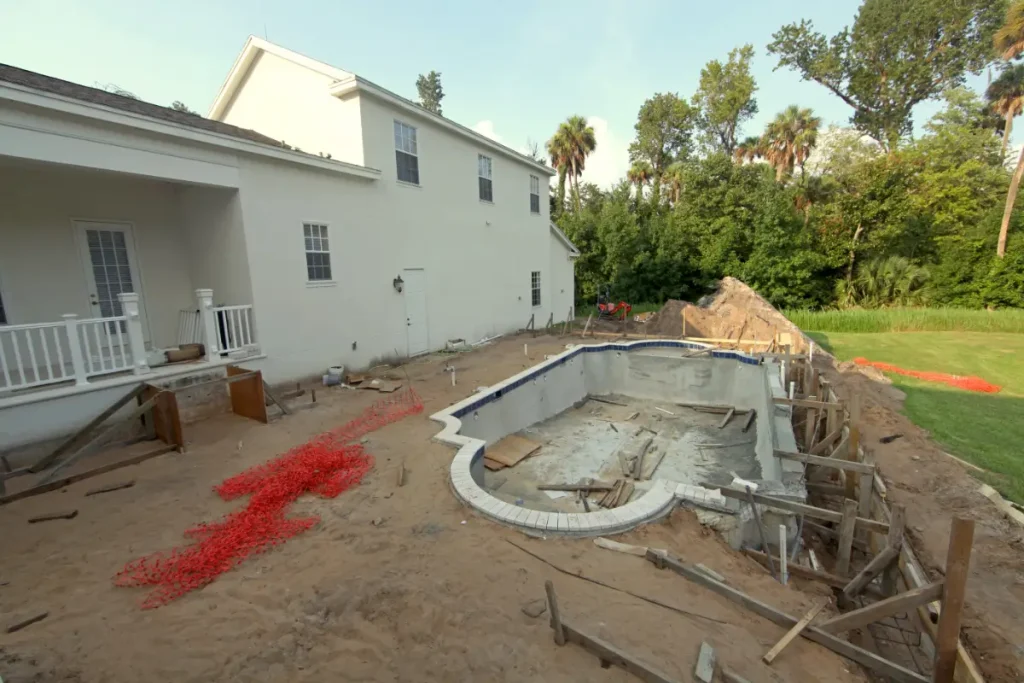 A large fiberglass pool size under construction, surrounded by dirt, construction materials, and a white two-story house.