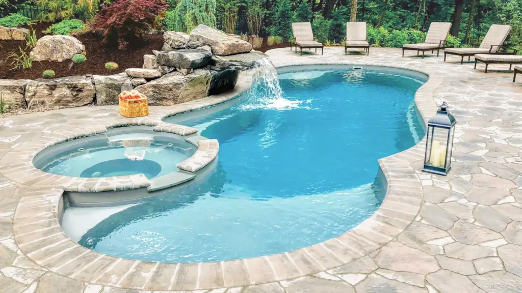 Backyard pool with integrated spa, cascading waterfall, and a stone-paved patio surrounded by lush greenery