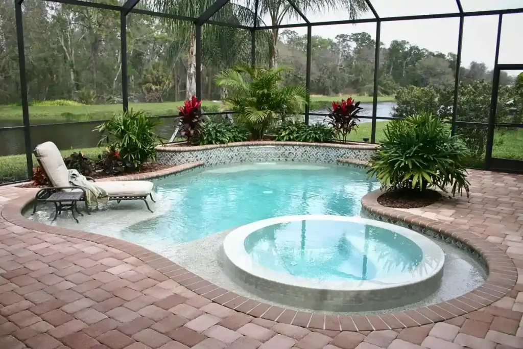 A stunning fiberglass pool with a built-in hot tub, surrounded by lush landscaping and an enclosed patio near a scenic lake.