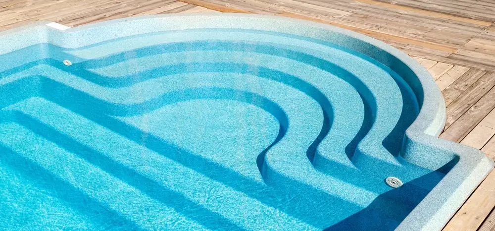 A sleek fiberglass pool kit featuring built-in curved steps and a wooden deck surrounding it.