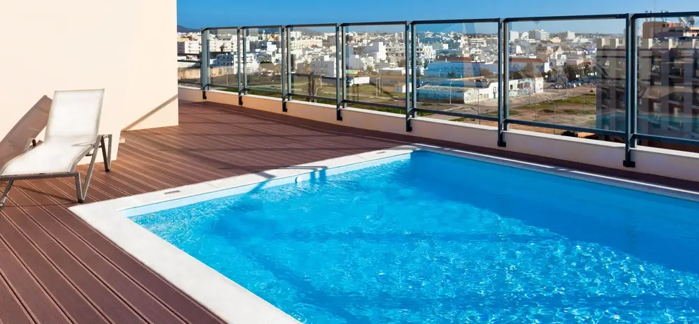 Rooftop fiberglass pool for sale with wooden decking, glass railings, and a cityscape view.