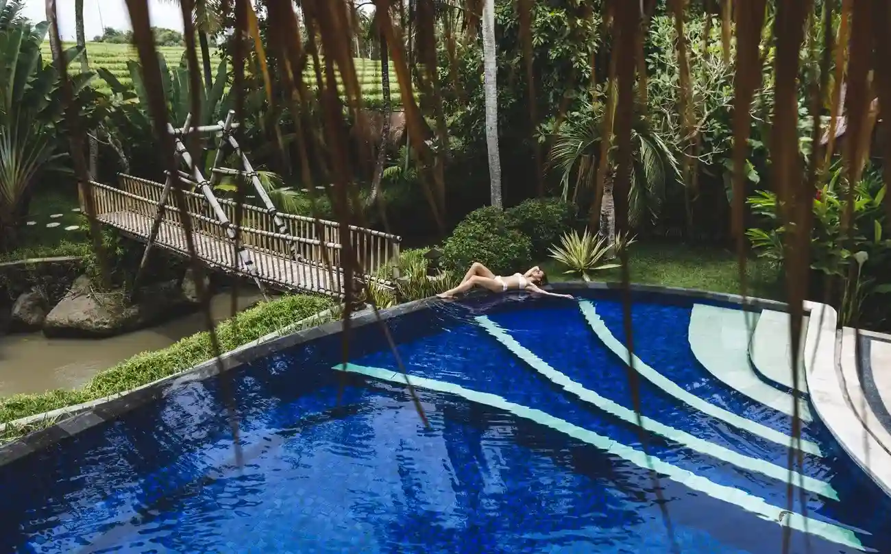 A vibrant fiberglass pool shell in a tropical setting, featuring bamboo bridges and lush greenery, with a woman relaxing by the water.