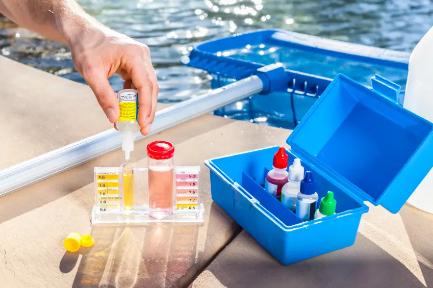 Hand using a pool water testing kit near a swimming pool to ensure water quality, with a blue cleaning kit box in view.