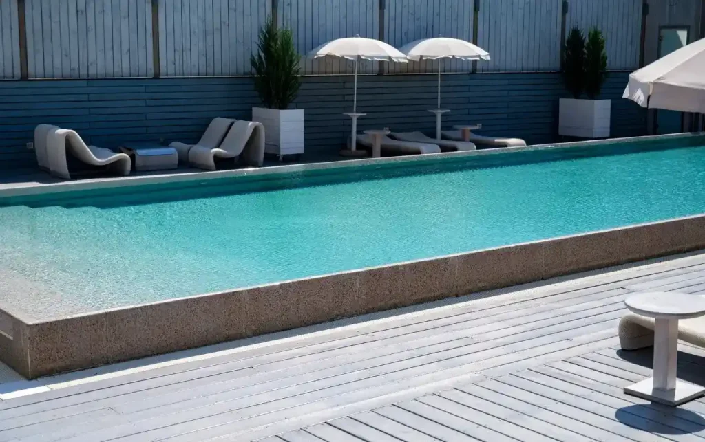 A sleek infinity-style concrete pool surrounded by a wooden deck and shaded lounge chairs.