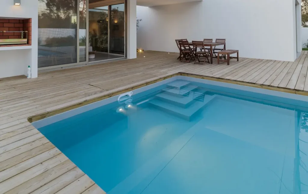 A modern fiberglass pool with a wooden deck and outdoor seating area in a stylish home setting.