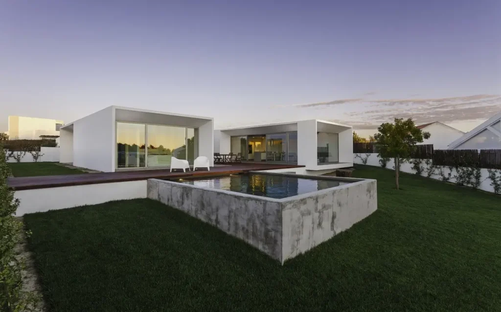 A modern semi above-ground pool with a sleek concrete finish in the backyard of a contemporary white house surrounded by a well-manicured lawn.