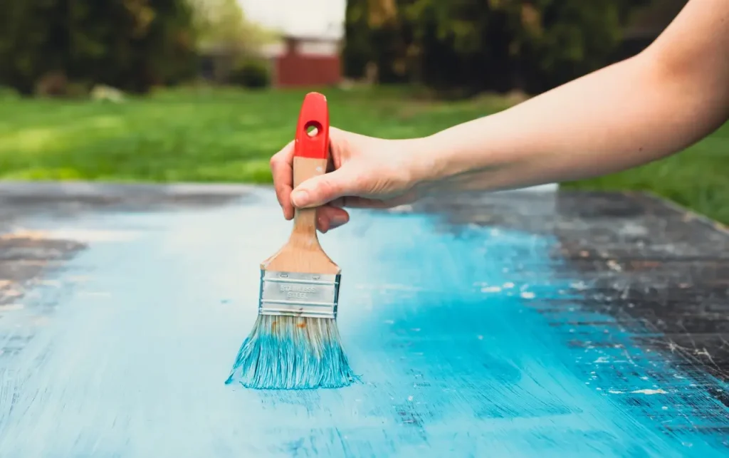 A hand holding a paintbrush applying blue concrete pool paint on a surface outdoors.