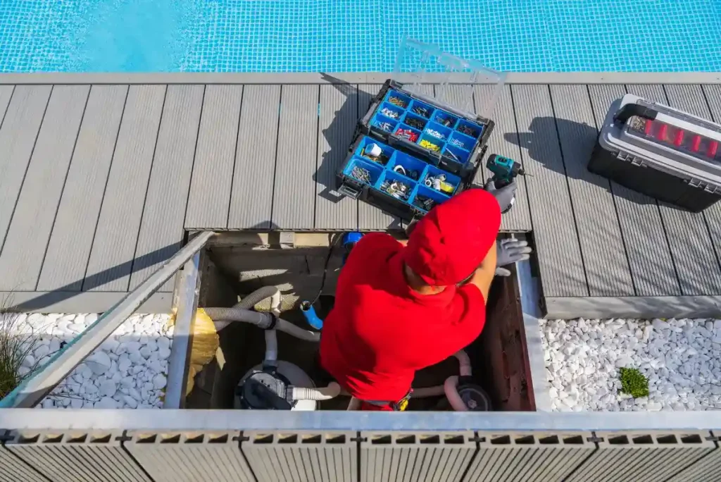 A pool maintenance worker in a red uniform servicing the filtration system of a pool with tools and equipment nearby.