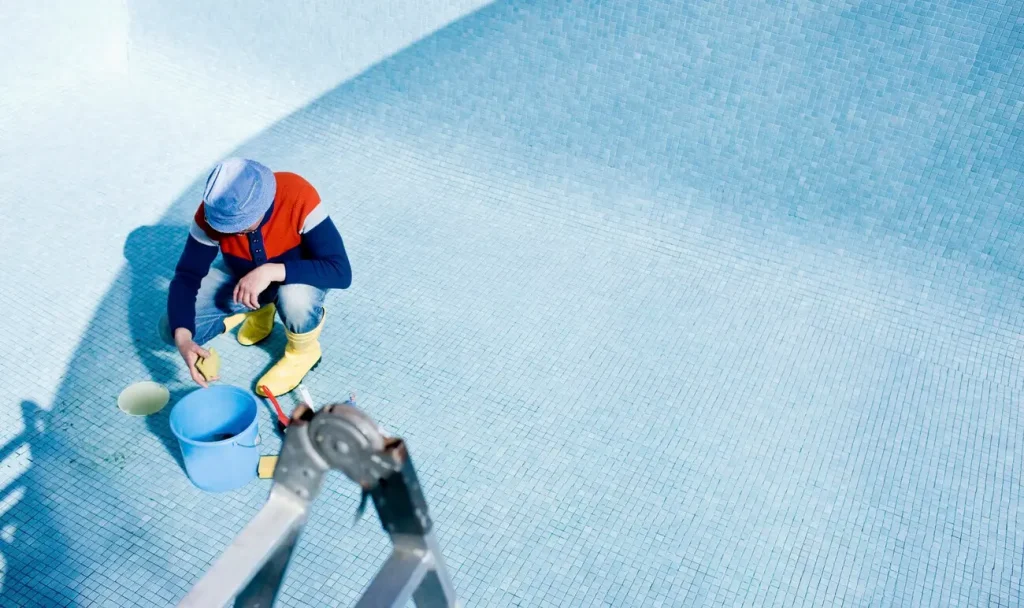 A person wearing yellow boots scrubbing the tiles of an empty above-ground swimming pool.