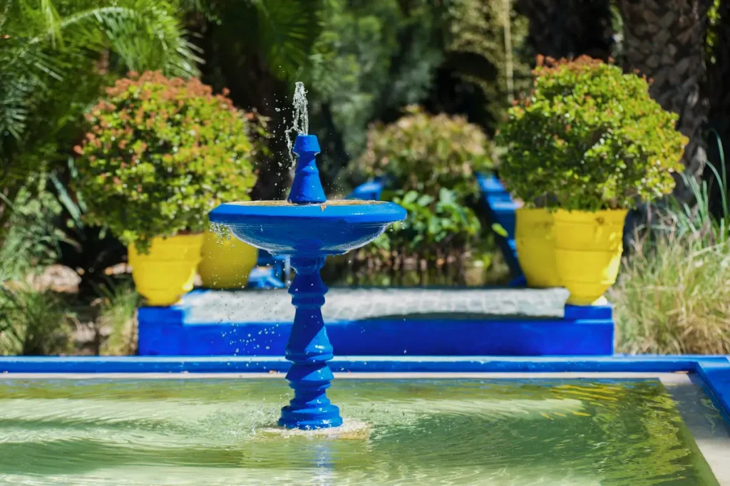 Above-ground pool with a vibrant blue fountain surrounded by lush greenery and yellow flower pots.