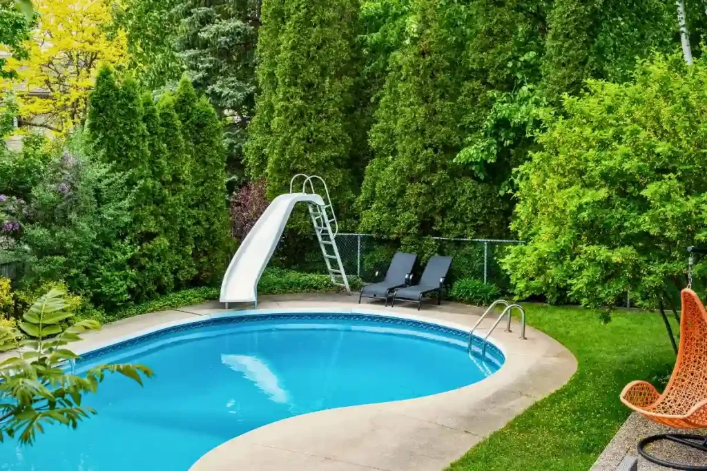 A DIY fiberglass pool featuring a water slide and relaxing lounge chairs, surrounded by greenery.