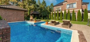 Luxurious rectangular pool installed by concrete pool companies near me, surrounded by a brick house and tall green trees.