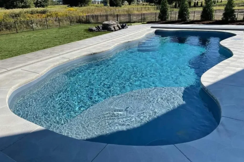 A modern fiberglass plunge pool with clear blue water, surrounded by a sleek light gray deck, a manicured lawn, and trees in the background.