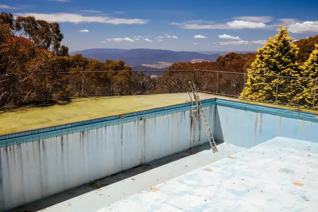 An empty outdoor swimming pool ready for pool remodeling, featuring peeling blue paint, a metal ladder, and a scenic mountainous backdrop surrounded by trees.