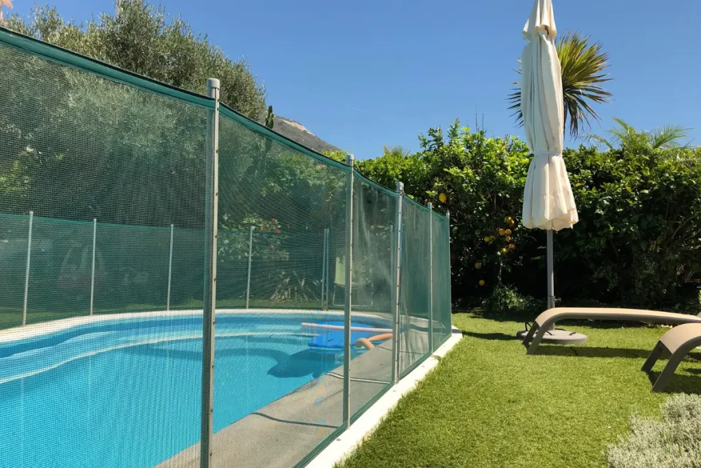 A backyard swimming pool enclosed by a safety fence, surrounded by lush green landscaping, a sun umbrella, and a lounge chair under a clear blue sky.