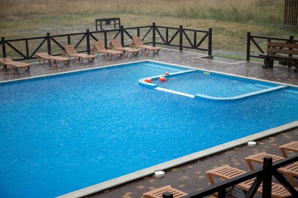 A custom-designed fiberglass pool with a unique shape, surrounded by lounge chairs and fencing during a rainy day near your location.